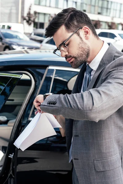 Geschäftsmann mit Wegnehmen von Lebensmitteln und Papieren in der Hand kontrolliert die Zeit, während er am Auto auf der Straße steht — Stockfoto