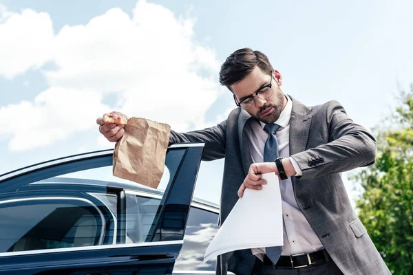 Businessman with take away food and papers in hands checking time while standing at car on street — Stock Photo