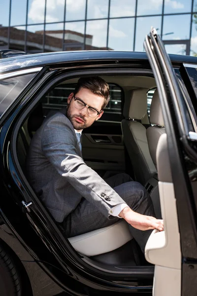 Вид сбоку застенчивого бизнесмена, отводящего взгляд, сидя на заднем сиденье с открытой дверью автомобиля — стоковое фото