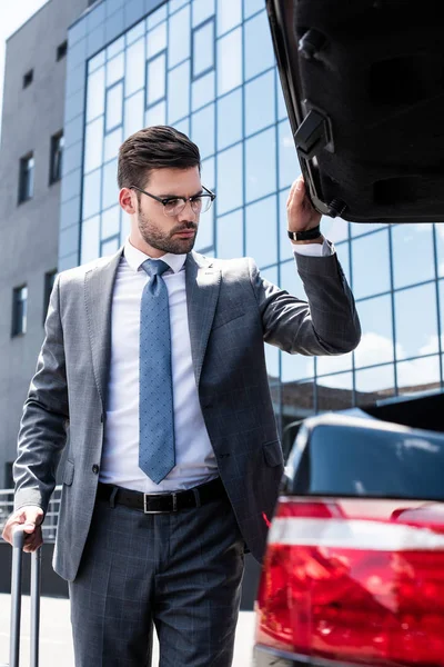 Young businessman in eyeglasses with wheeled bag opening car trunk at street — Stock Photo