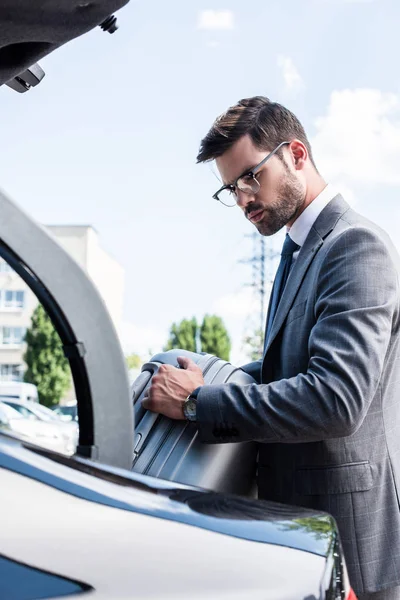 Hombre de negocios guapo en gafas que ponen la bolsa de ruedas en el maletero del coche en la calle - foto de stock