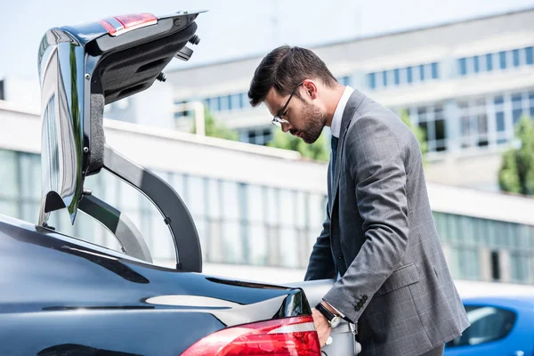 Side view of businessman in eyeglasses putting wheeled bag in car trunk at street — Stock Photo