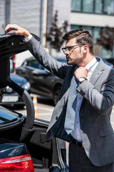 Young businessman in eyeglasses closing car trunk at street — Stock Photo