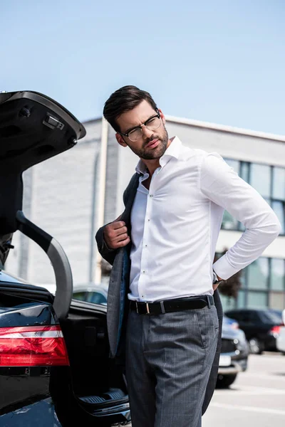 Vista de ángulo bajo del hombre de negocios guapo quitándose la chaqueta y mirando hacia otro lado cerca del coche - foto de stock