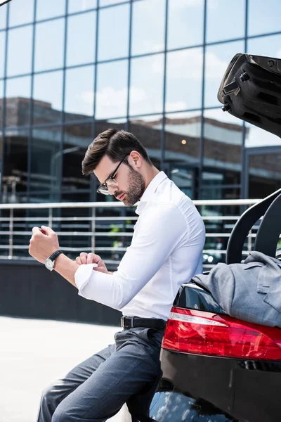 Vista laterale dell'uomo d'affari in occhiali che rotola maniche di camicia bianca mentre si siede sul tronco dell'auto in strada — Foto stock