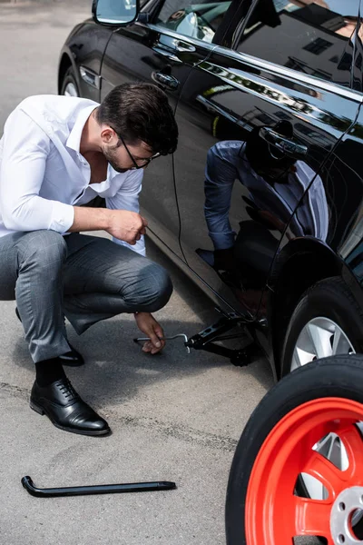 Jovem empresário sentado perto de carro quebrado e usando parafuso jack na rua — Fotografia de Stock