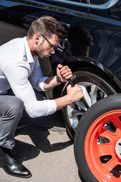 Concentrated young businessman in eyeglasses using wheel spanner for wheel replacement at street — Stock Photo