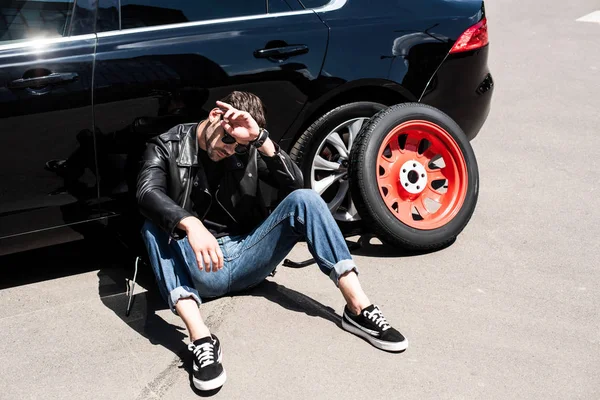 Stylish tired man in sunglasses wiping forehead near tools, tire and broken car at street — Stock Photo