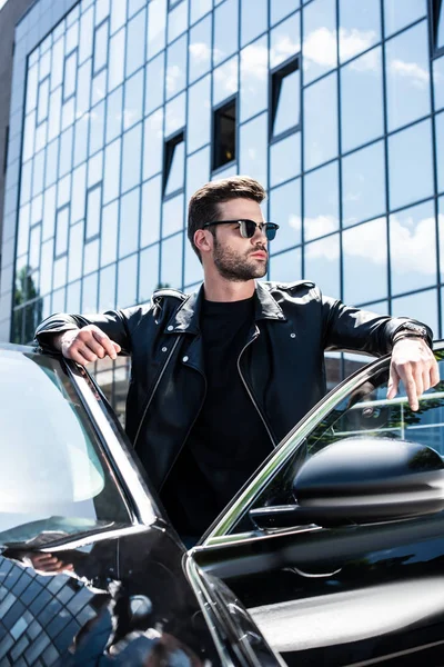 Hombre elegante guapo en chaqueta de cuero y gafas de sol posando cerca de coche en la calle - foto de stock