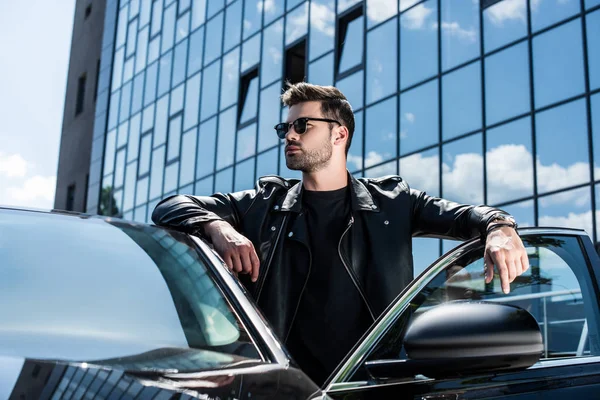 Молодой стильный мужчина в кожаной куртке и солнцезащитных очках глядя в сторону автомобиля на улице — стоковое фото