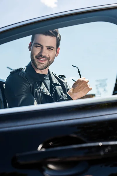 Jovem sorridente em jaqueta de couro segurando óculos de sol perto de seu carro na rua — Fotografia de Stock