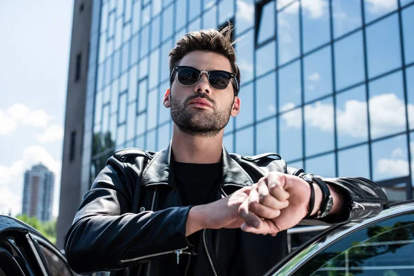 Low angle view of stylish man in leather jacket and sunglasses touching wristwatch near car — Stock Photo