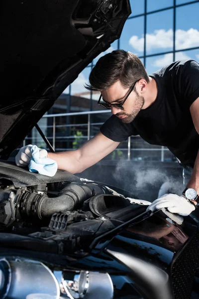 Handsome young man in working gloves holding rag and looking at engine of broken car with smoke coming out — Stock Photo