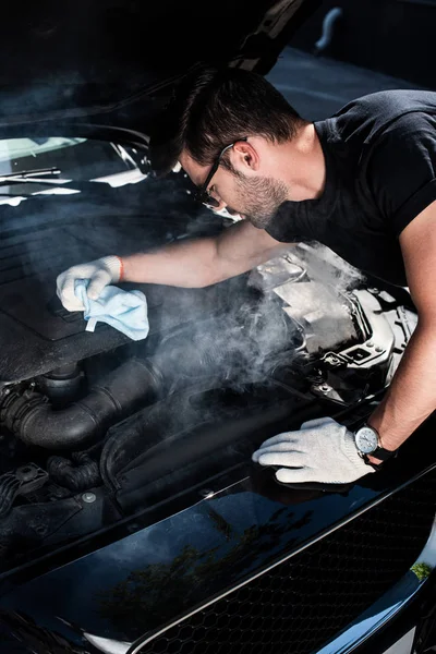 Seitenansicht eines jungen Mannes in Arbeitshandschuhen, der Lappen hält und den Motor eines kaputten Autos betrachtet, aus dem Rauch aufsteigt — Stockfoto