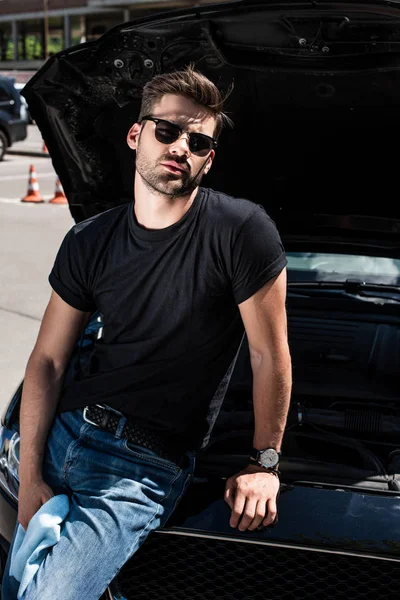 Schöner junger Mann mit Sonnenbrille und Lappen neben Auto mit geöffneter Motorhaube auf der Straße — Stockfoto