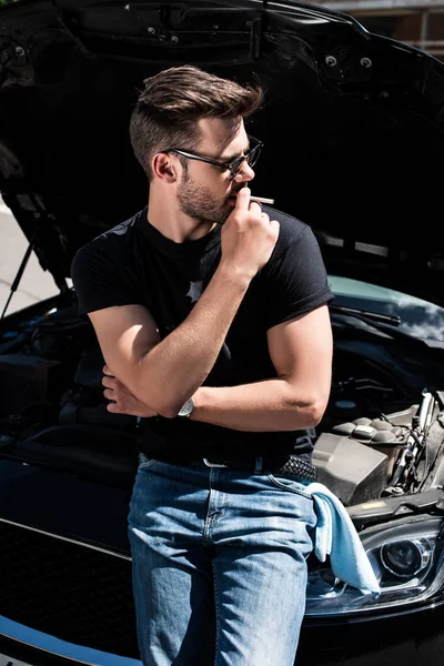 Side view of stylish man in sunglasses smoking cigarette near car with opened bonnet — Stock Photo