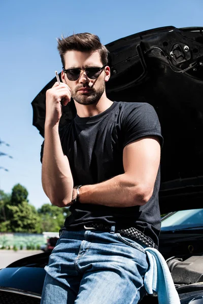 Молодой стильный мужчина в солнечных очках курит сигарету рядом со сломанной машиной с открытым капотом — стоковое фото