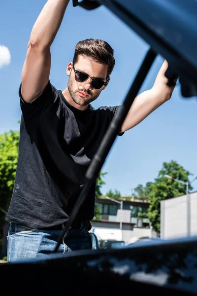 Low angle view of young man in sunglasses opening bonnet at street — Stock Photo