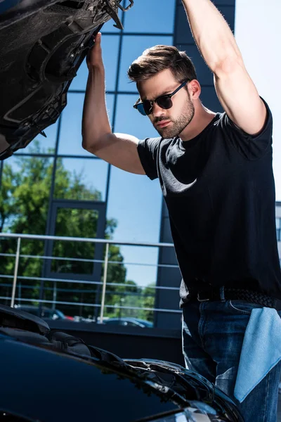 Низкий угол зрения молодого человека в солнцезащитных очках, проверяющего двигатель автомобиля на улице — стоковое фото