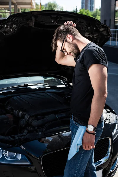 Side view of confused man scratching head near broken car engine at street — Stock Photo