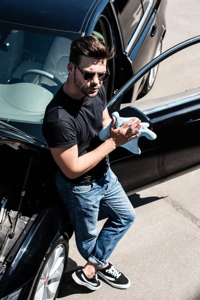 Vista de ángulo alto de hombre joven en gafas de sol limpiando las manos por trapo cerca del coche con el capó abierto - foto de stock