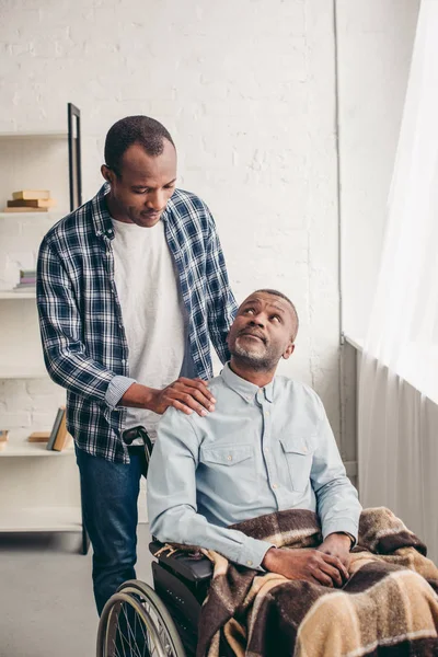 Erwachsener afrikanisch-amerikanischer Sohn schaut behinderten Vater im Rollstuhl an — Stockfoto