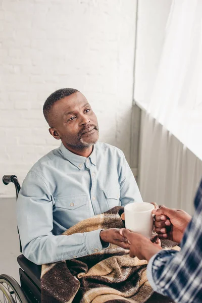 Cropped shot of adult son giving cup of hot beverage to disabled senior father — Stock Photo