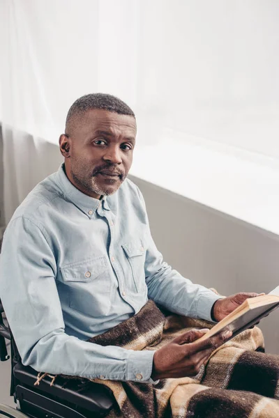 Visão de alto ângulo de deficiente homem idoso em cadeira de rodas segurando livro e olhando para a câmera — Fotografia de Stock