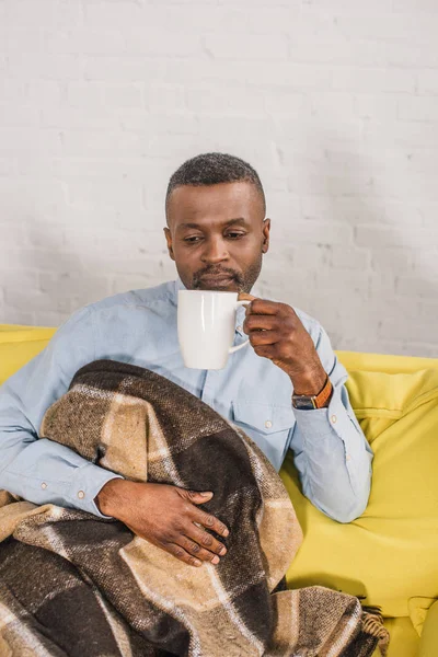 Старший афроамериканський чоловік п'є чай, сидячи на дивані з пледом — стокове фото