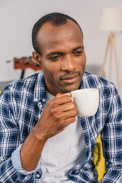 Pensativo joven afroamericano hombre sosteniendo taza de café en casa - foto de stock