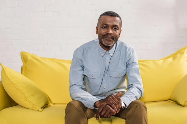 Senior homme afro-américain assis sur le canapé jaune et souriant à la caméra — Photo de stock