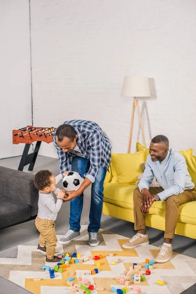 Glücklicher Vater und Großvater beim Anblick eines entzückenden Kleinkindes, das aus einer Babyflasche trinkt, während es zu Hause mit Spielzeug spielt — Stockfoto