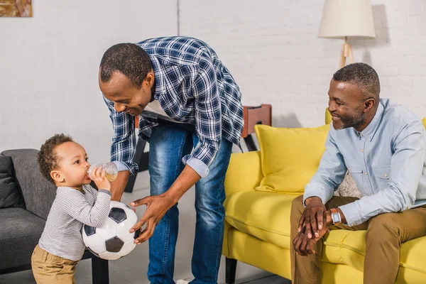 Счастливый отец и дедушка смотрят на очаровательного малыша, который держит футбольный мяч и пьет из детской бутылочки дома — стоковое фото