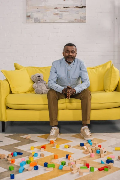Hombre afroamericano senior mirando a la cámara mientras está sentado en el sofá amarillo en la habitación con bloques de colores en la alfombra - foto de stock