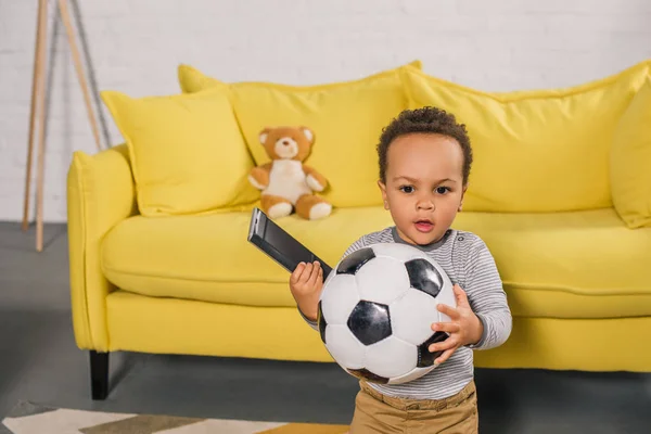 Adorable african american toddler holding soccer ball and remote controller at home — Stock Photo