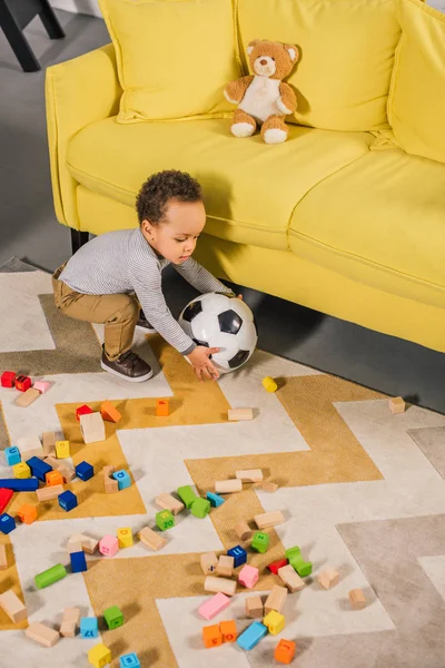 Visão de alto ângulo de criança pequena bonito brincando com bola de futebol e blocos coloridos em casa — Fotografia de Stock