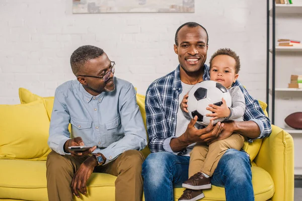 Feliz hombre mayor sosteniendo mando a distancia y mirando sonriente joven padre e hijo sentado en el sofá con pelota de fútbol - foto de stock