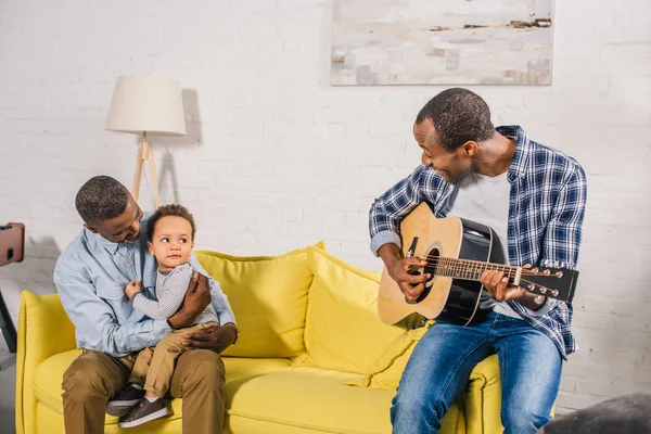Smiling young man playing guitar and looking at happy grandfather and grandson at home — Stock Photo