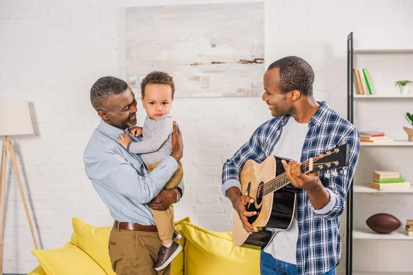 Smiling young man playing guitar and looking at happy grandfather holding grandson at home — Stock Photo