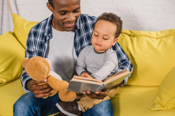 Cropped shot of smiling father holding teddy bear and reading book to little son — Stock Photo