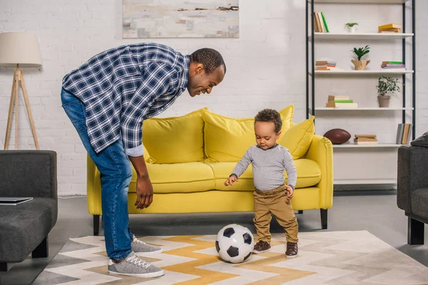 Feliz jovem pai olhando para o pequeno filho sorrindo jogando com bola de futebol em casa — Fotografia de Stock