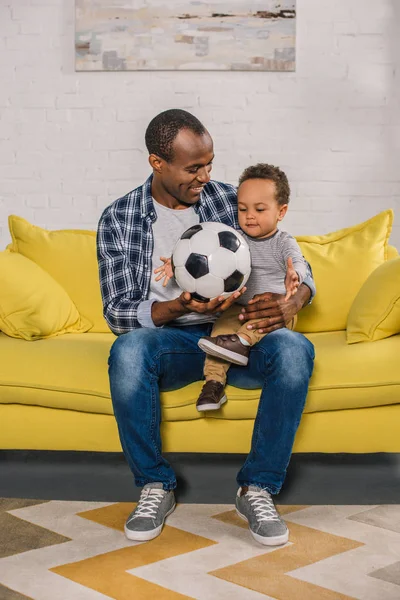 Glücklicher junger Vater hält Fußballball in der Hand und schaut seinen süß lächelnden Sohn an — Stockfoto