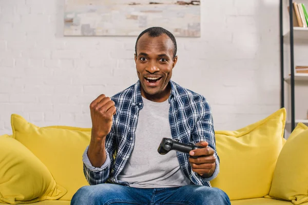 Alegre africano americano hombre sonriendo a cámara mientras jugando con joystick en casa - foto de stock