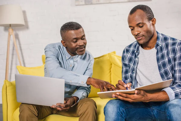 Senior man using laptop and looking at adult son taking notes in notepad at home — Stock Photo