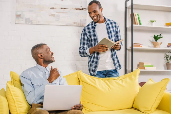 Senior man with laptop and adult son with book smiling each other at home — Stock Photo