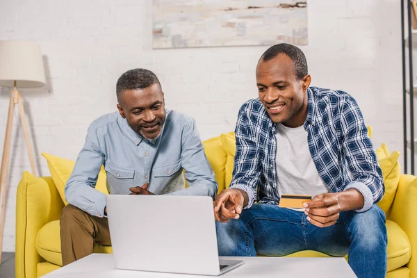 Smiling senior man and adult son with credit card using laptop together at home — Stock Photo