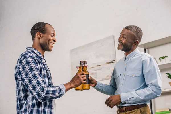 Pai sênior feliz e filho adulto batendo garrafas de cerveja e sorrindo uns aos outros em casa — Fotografia de Stock