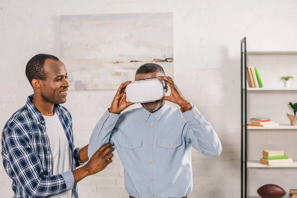 Sorridente giovane uomo guardando padre anziano utilizzando auricolare realtà virtuale a casa — Foto stock