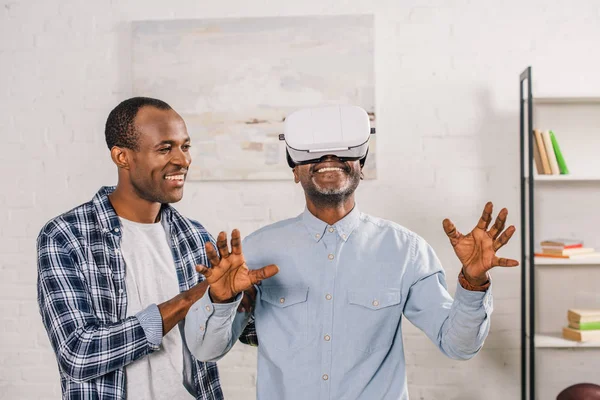 Smiling young man looking at happy senior father using virtual reality headset at home — Stock Photo