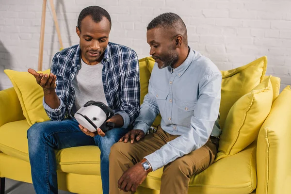 Homme âgé souriant avec fils adulte utilisant un casque de réalité virtuelle à la maison — Photo de stock